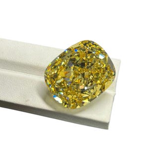 May include: A large, faceted yellow diamond with a cushion cut. The gemstone is set on a white surface, showcasing its brilliant facets and golden hue. The diamond's shape is a modified cushion, with rounded corners and a rectangular body.