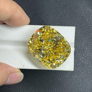 May include: A large, cushion-cut yellow gemstone with many facets, reflecting light. The gemstone is held on a white surface, with a dark background. The cut of the stone is rectangular with rounded corners.