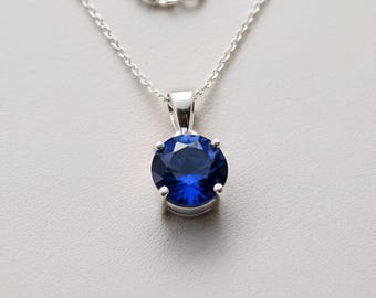 May include: A silver necklace featuring a round, deep blue gemstone pendant. The pendant is held in place by four prongs, and the chain is a delicate silver. The gemstone has a faceted surface that reflects light.