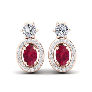 Könnte beinhalten: Ein Paar roségoldene Ohrringe. Jeder Ohrring hat einen zentralen, oval geschliffenen, tiefroten Edelstein, der von einem Kranz aus kleinen, klaren Edelsteinen umgeben ist. Ein runder, klarer Edelstein sitzt auf jedem Ohrring.