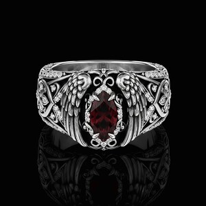 May include: A silver ring with an ornate design featuring angel wings and a central, teardrop-shaped, red gemstone. The ring is encrusted with small, clear gemstones and has a detailed, textured band. The ring is displayed against a black background.