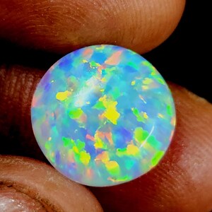 May include: A spherical opal gemstone with a play of colour. The opal features a mix of blue, yellow, green, and orange hues, set against a milky white background. The gemstone is held between fingers, highlighting its polished surface.