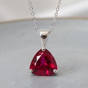 Może przedstawiać: Srebrny naszyjnik z trójkątną, fasetowaną, ciemnoczerwoną zawieszką z kamienia szlachetnego. Zawieszka zawieszona jest na delikatnym łańcuszku. Kamień szlachetny ma brylantowy szlif, odbijający światło. Naszyjnik jest prezentowany na neutralnym tle.