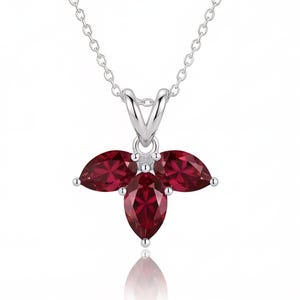 May include: A silver necklace featuring a pendant with three marquise-cut deep red gemstones arranged in a floral-like cluster. The pendant is suspended from a delicate silver chain, showcasing a polished metallic finish and faceted stone surfaces.