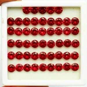 May include: A selection of round, faceted, deep red gemstones displayed in a white tray. The gems are neatly arranged, highlighting their brilliant cut and rich colour. The tray is held, implying a presentation or storage setting.
