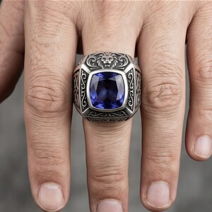 Puede incluir: Un anillo plateado con una gran gema cuadrada de color azul oscuro. El anillo presenta detalles ornamentados, incluyendo una cabeza de león y volutas. El anillo se lleva en un dedo.