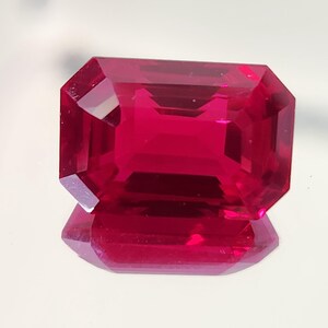 Puede incluir: Una piedra preciosa de rubí tallada en forma rectangular. La gema es de un color rojo intenso y vibrante, con bordes afilados y limpios y una superficie reflectante. La piedra se muestra sobre una superficie reflectante, creando una imagen especular.