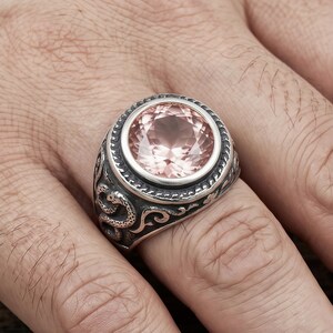 Peut inclure: Bague en argent ornée d'une grosse pierre précieuse rose pâle et ronde. La bague présente un motif orné avec un serpent et des volutes sur le bandeau. La pierre est sertie dans un sertissage à lunette.