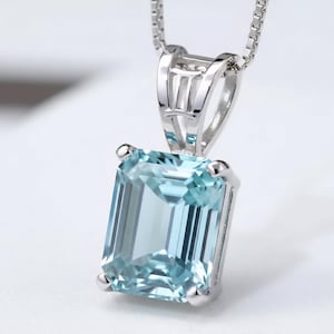 Puede incluir: Un collar con colgante de plata con una piedra preciosa rectangular de color azul claro. La piedra está engastada en un bisel de plata y cuelga de una fina cadena de plata. El colgante tiene un diseño clásico y elegante.