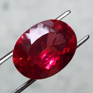 Puede incluir: Una gema facetada, de forma ovalada, de color rojo intenso, sujeta con pinzas plateadas. La superficie de la gema refleja la luz, mostrando su talla y claridad. La gema es de un color rojo intenso y saturado, con un aspecto pulido.