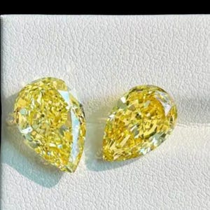 May include: Two pear-shaped, fancy vivid yellow diamonds. The gems are set against a white textured background. The diamonds have multiple facets that reflect light, creating a brilliant sparkle. The overall theme is luxury and fine jewelry.