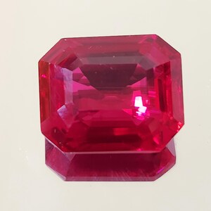 Puede incluir: Una gema roja intensa, tallada en forma rectangular. La gema tiene muchas facetas que reflejan la luz. La piedra está sobre un fondo claro, con su reflejo visible debajo. Una pieza de joyería elegante.