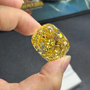 May include: A large, cushion-cut yellow diamond. The gemstone is held between fingers, showcasing its brilliant facets and intense colour. The diamond's shape is rectangular with rounded corners, reflecting light in a dazzling display.