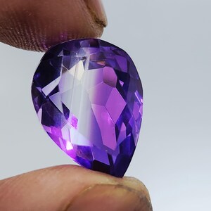 Puede incluir: Una gema de amatista facetada en forma de pera. La gema muestra un degradado de tonos púrpuras, que van del violeta oscuro a tonos más claros. El corte revela múltiples facetas, realzando su brillo y claridad. Sostenida entre los dedos sobre un fondo neutro.