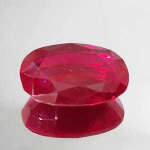 Puede incluir: Una gema de rubí facetada de forma ovalada. El rubí es de un color rojo intenso y vibrante con una superficie pulida. El reflejo de la gema es visible debajo, creando un efecto de espejo. La gema es un símbolo de amor y pasión.