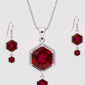 May include: A silver necklace and earring set featuring hexagonal red gemstones. The pendant and earrings have a geometric design with a larger gemstone at the top and a smaller one below the pendant and earrings. The jewelry is set against a white background.