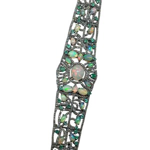 Puede incluir: Una pulsera plateada con un diseño calado, con una gran ópalo central y ópalos y esmeraldas más pequeños. La pulsera está adornada con pequeños acentos brillantes.