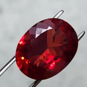 May include: A deep red, oval-cut gemstone, held by silver tweezers. The facets of the gem catch the light, creating a play of internal reflections. The colour varies from a dark, rich red to brighter highlights, showcasing its polished surface.