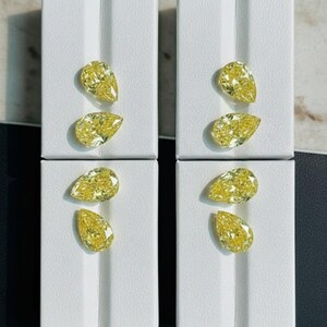 May include: Four sets of two pear-shaped yellow diamonds displayed on white rectangular platforms. The diamonds have a vibrant yellow hue and are presented in a symmetrical arrangement, showcasing their cut and clarity. The background is a neutral tone.