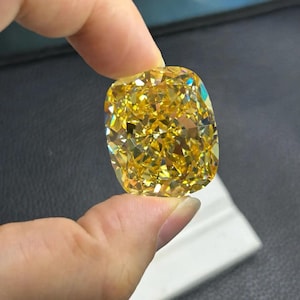May include: A large, cushion-cut, yellow diamond held between fingers. The gemstone has a brilliant, multifaceted surface that reflects light, showcasing its vibrant colour and clarity. The diamond's shape is square with rounded corners.