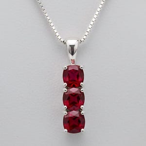 Può includere: Una collana con pendente in argento con tre pietre preziose ovali rosso scuro disposte verticalmente. Il pendente è sospeso a una sottile catena d'argento, offrendo un aspetto classico ed elegante. Le pietre sono tenute da griffe d'argento.