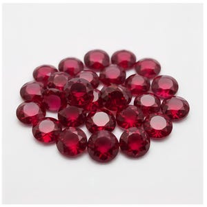 Könnte beinhalten: Eine Ansammlung kleiner, runder, facettierter roter Edelsteine. Jeder Stein weist einen Brillantschliff mit mehreren dreieckigen Facetten auf, die das Licht reflektieren und eine tiefrote Farbe sowie ein funkelndes Aussehen der losen Steine erzeugen.