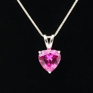 Pode incluir: Um colar de prata com um pingente de pedra preciosa rosa em forma de coração. O pingente é de um rosa vibrante e é fixado em uma moldura de prata. A corrente do colar é uma corrente de prata delicada.