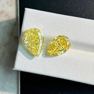 May include: Two pear-shaped, fancy yellow diamonds are displayed on a white surface. The diamonds are cut to maximize brilliance, reflecting light. The gems are a vibrant yellow color, showcasing their clarity and quality. These diamonds are suitable for jewelry.