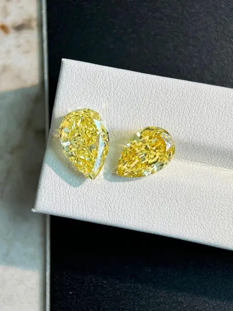 May include: Two pear-shaped, fancy yellow diamonds are displayed on a white surface. The diamonds are cut to maximize brilliance, reflecting light. The gems are a vibrant yellow color, showcasing their clarity and quality. These diamonds are suitable for jewelry.
