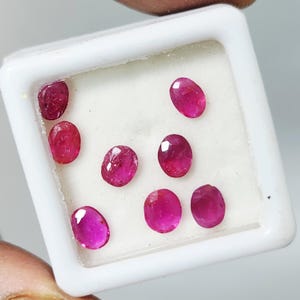 Puede incluir: Una colección de gemas rojas vibrantes, de corte ovalado, exhibidas en un contenedor cuadrado blanco. Las gemas están dispuestas sobre una superficie blanca, mostrando sus superficies pulidas y su rico color. El contenedor es sostenido por una mano.