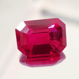 Puede incluir: Una gema de rubí rectangular y facetada, de color rojo intenso. La gema está tallada con múltiples facetas, reflejando la luz y creando un brillo brillante. El rubí está sobre un fondo blanco, con su reflejo visible debajo.