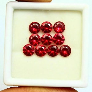 May include: Nine round, faceted, deep red gemstones are arranged in a white tray. The gems have a brilliant cut, reflecting light. The tray is held by a hand. These gemstones are suitable for jewelry making.