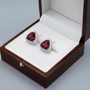 May include: A pair of stud earrings featuring triangular-cut deep red gemstones surrounded by a halo of small clear stones set in silver-toned metal. The earrings are displayed inside a dark brown wooden box with a white interior lining.