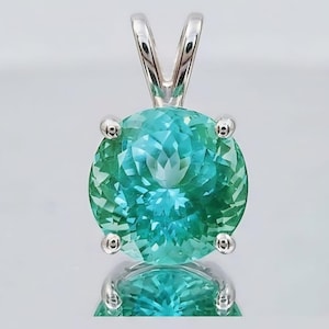 May include: A round, faceted pendant with a vibrant teal and green gemstone. The gemstone is held in place by four silver prongs, and a silver bail is attached at the top. The pendant is displayed against a reflective surface.