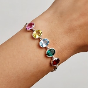 May include: A silver bracelet with six oval gemstones in a row. The gemstones are pink, yellow, light blue, green, and red. The bracelet is connected by a delicate chain.