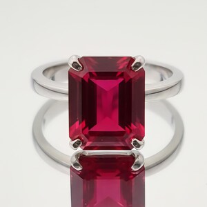 Op de afbeelding: Een zilveren ring met een grote, rechthoekige, dieprode edelsteen. De edelsteen wordt op zijn plaats gehouden door vier klauwen, en de ringband is van gepolijst zilver. De ring wordt getoond op een reflecterend oppervlak.