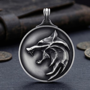 May include: A silver pendant with a detailed wolf head design inside a circular frame. The wolf is shown with its mouth open, teeth bared. The pendant has a bail for a chain and is set against a dark backdrop.