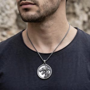 May include: Silver pendant necklace with a wolf head emblem. The circular pendant hangs from a chain. The person in the image is wearing a black t-shirt.