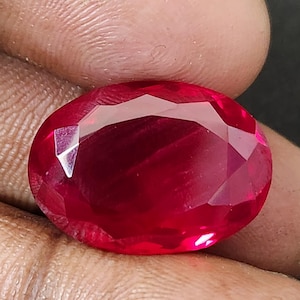 Puede incluir: Una piedra preciosa de rubí facetada, de forma ovalada. La gema es de un color rojo intenso y saturado con múltiples facetas que reflejan la luz. La piedra preciosa se sostiene en una mano, mostrando su tamaño y brillo.