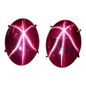 Op de afbeelding: Twee ovale, dieprode edelstenen met een stervormig patroon van witte lijnen. De edelstenen zijn gezet met kleine, donkere metalen pinnen. De stenen hebben een gepolijst, reflecterend oppervlak.