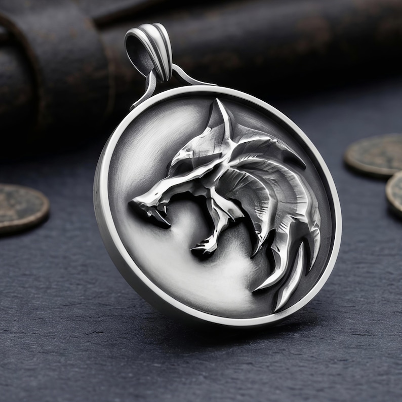 May include: A silver pendant with a detailed wolf head design. The round pendant has a raised relief of a snarling wolf's face. It has a loop at the top for a chain. The background is dark.