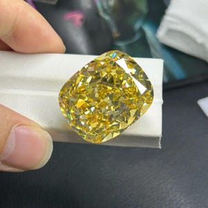 May include: A large, cushion-cut yellow diamond with many facets. The gemstone is a vibrant yellow colour and is displayed on a white surface. The diamond's cut reflects light, creating a sparkling effect.