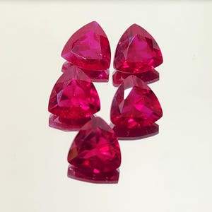 May include: Five triangular-cut, faceted gemstones in a vibrant, deep red hue. The gems are arranged on a reflective surface, showcasing their polished facets and internal brilliance. The gems are likely rubies, known for their rich color and clarity.