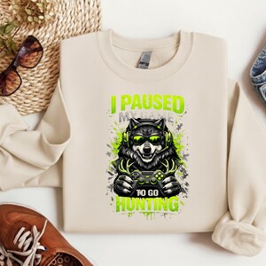 May include: Beige sweatshirt with a graphic of a wolf wearing headphones and sunglasses, holding a game controller. The text reads "I PAUSED MY GAME TO GO HUNTING" in green. The wolf is surrounded by green and gray splatter.
