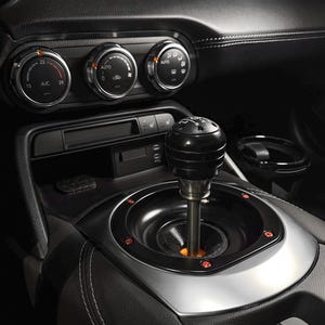 Può includere: Primo piano degli interni di un'auto con un pomello del cambio nero e una base argentata. Il soffietto del cambio è nero con luci rosse di accento. Il cruscotto include quadranti per il controllo del clima e un portabicchieri.