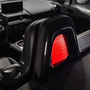 Może przedstawiać: Zbliżenie czarnego zagłówka fotela samochodowego z błyszczącym wykończeniem. Zagłówek posiada jaskrawoczerwony, teksturowany panel. W tle widoczne jest wnętrze samochodu, w tym kierownica i deska rozdzielcza.