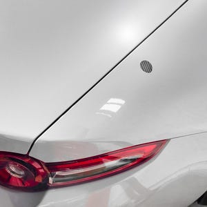 May include: Close-up of a white sports car's rear section, showcasing a red taillight and a small, circular vent. The car's body has a glossy finish, with a black accent line and a partially visible tire.
