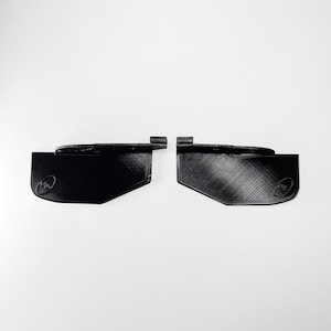Puede incluir: Dos componentes de plástico negro impresos en 3D con un cuerpo plano y rectangular y un mecanismo de bisagra que sobresale en el borde superior. Cada pieza muestra un sutil patrón de textura entrecruzada y un pequeño logotipo circular en relieve.