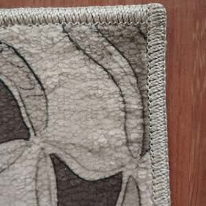 May include: Detail of a rug with a floral design in neutral tones. The rug's pattern includes shades of grey and brown. It has a stitched border and is placed on a wooden surface. The overall aesthetic is muted and textured.