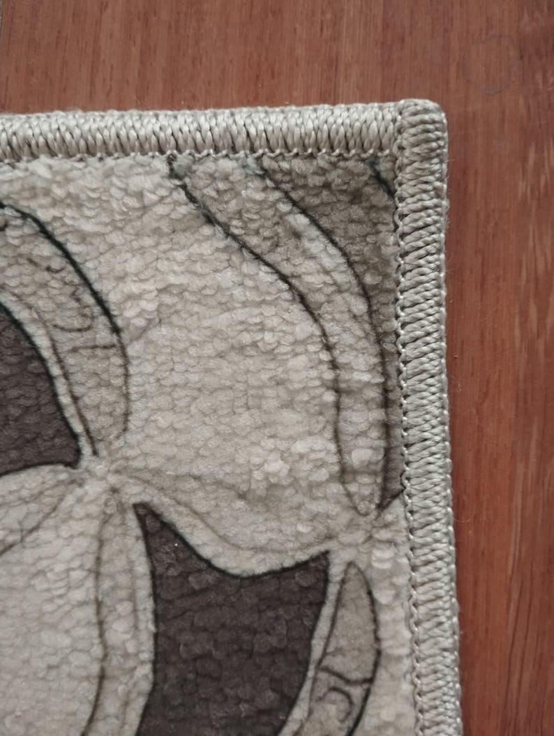 May include: Detail of a rug with a floral design in neutral tones. The rug's pattern includes shades of grey and brown. It has a stitched border and is placed on a wooden surface. The overall aesthetic is muted and textured.